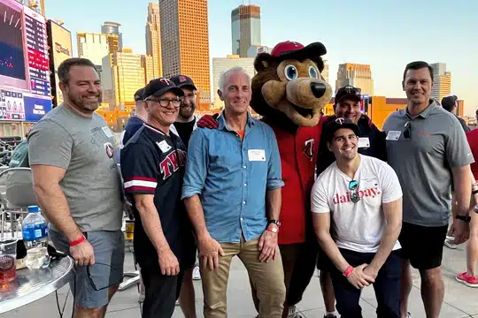 A group of seven smiling men and a mascot stand together on a rooftop with a city skyline in the background. They appear to be at an event, wearing casual attire and name tags.