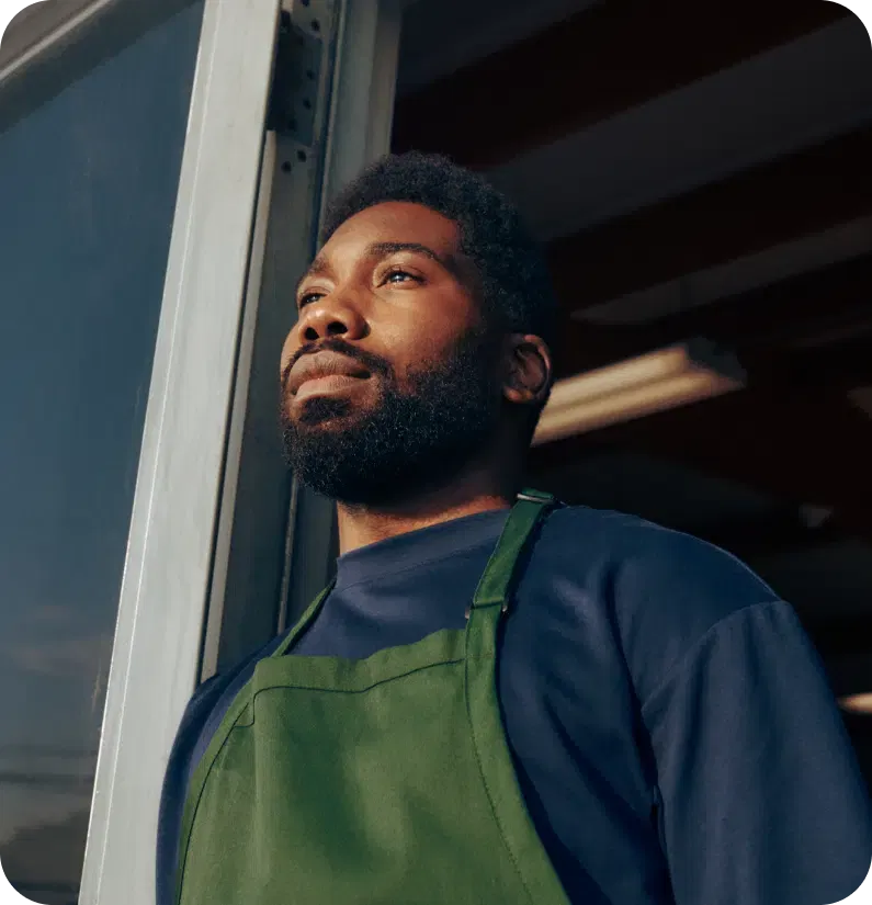 A person in a green apron stands next to a doorway, looking in the distance with a thoughtful expression.