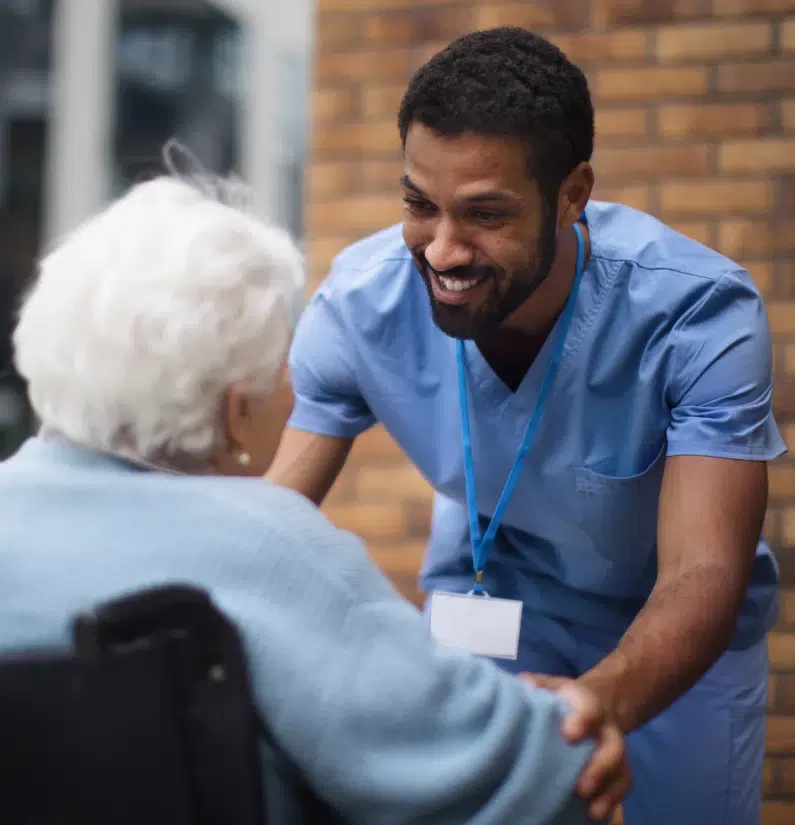 A healthcare professional wearing blue scrubs and a name badge smiles and holds hands with an elderly person seated in a wheelchair.