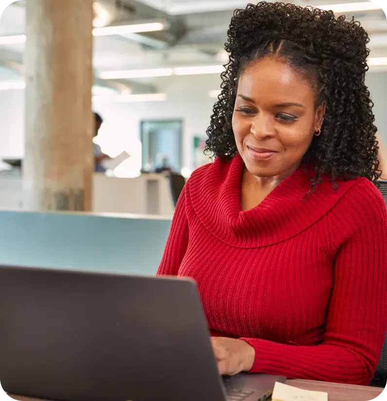 A woman with curly hair wearing a red sweater works on a laptop in a modern office setting.
