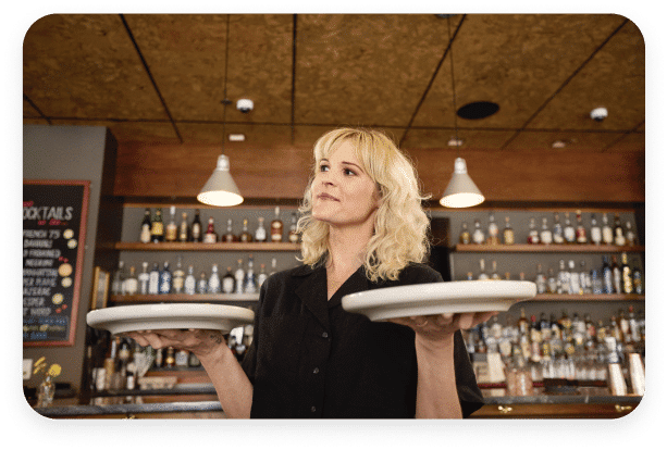 A woman with blonde hair is standing in a bar, holding two empty white plates, one in each hand. She is wearing a black shirt. Behind her, a well-stocked bar glows under several hanging lights, reminiscent of celebrations where spot bonuses reward hardworking employees.