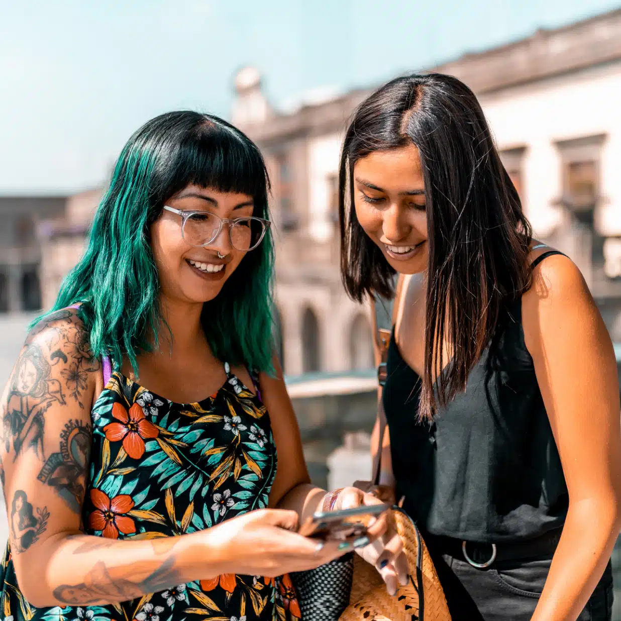 Two women stand together outside, one with green hair and tattoos, both smiling as they look at a smartphone.