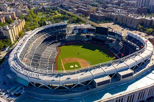 An aerial view of a baseball stadium with an empty field and stands, surrounded by urban buildings and greenery.